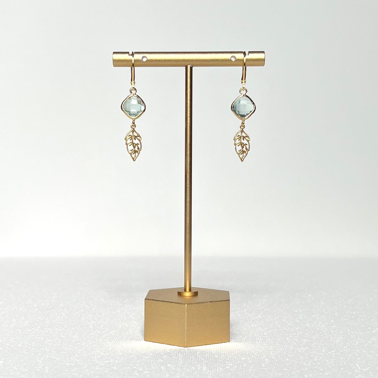 This earring set features a pale green glass charm in a gold bezel with a gold leaf, hung on a French lever back style hook. 