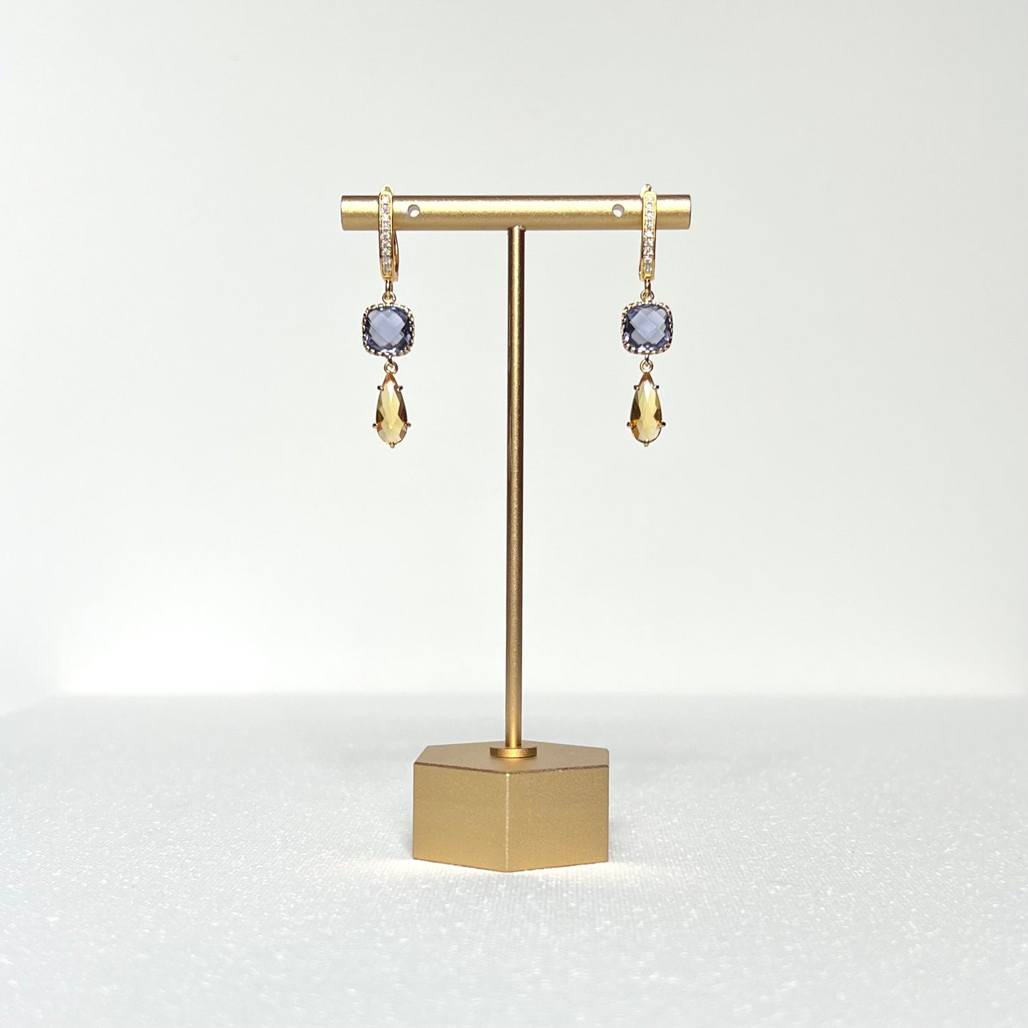 This pair of earrings features a tanzanite colored glass charm in a fancy prong bezel with a citrine colored glass teardrop shaped prong set charm hung by a CZ crested gold filled post style lever back earring. 